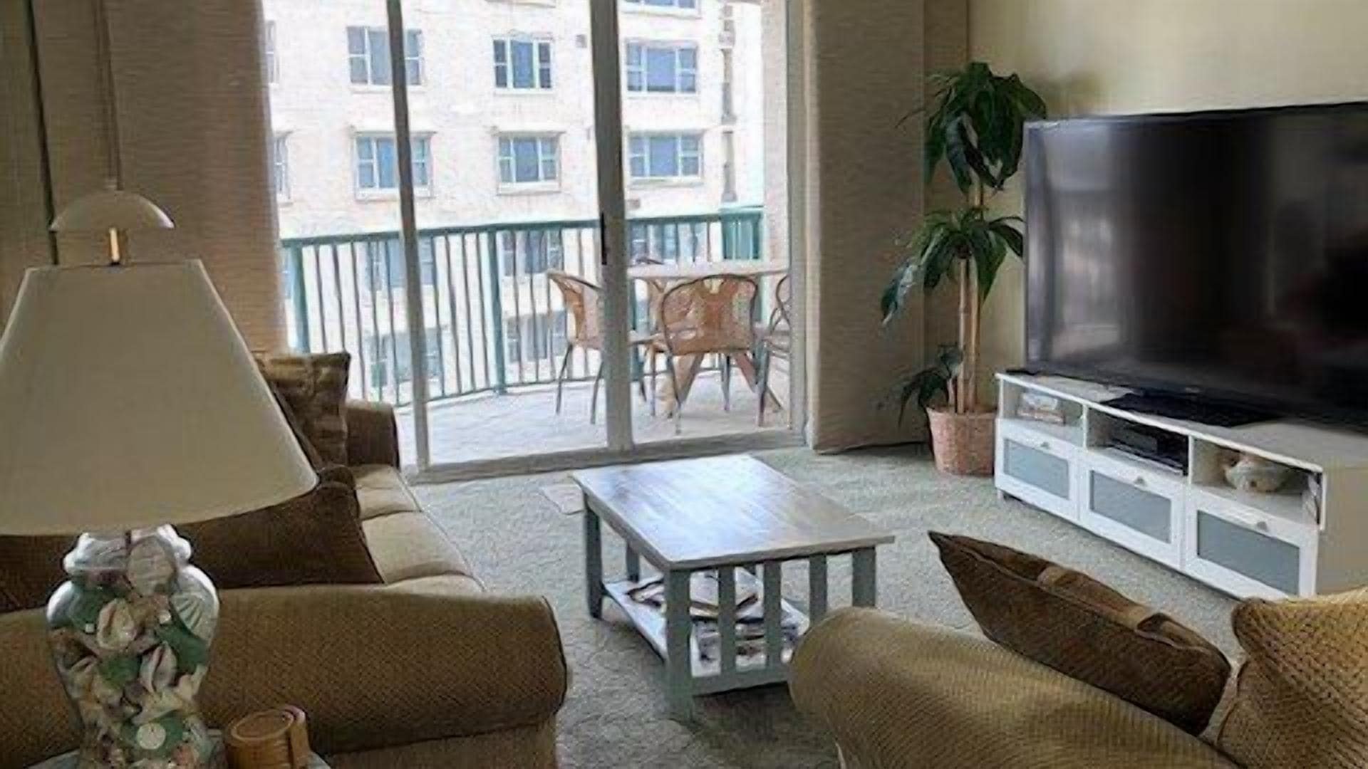 Una sala de estar con un sofá, una mesa de centro, un televisor sobre un mueble bajo y un balcón con vistas a edificios.