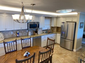 A modern kitchen with white cabinets, stainless steel appliances, and a dining area with wooden chairs. - #705 Sandcastle 2/2 Oceanfront CondoTraffic Free Beach (Ponce Inlet)