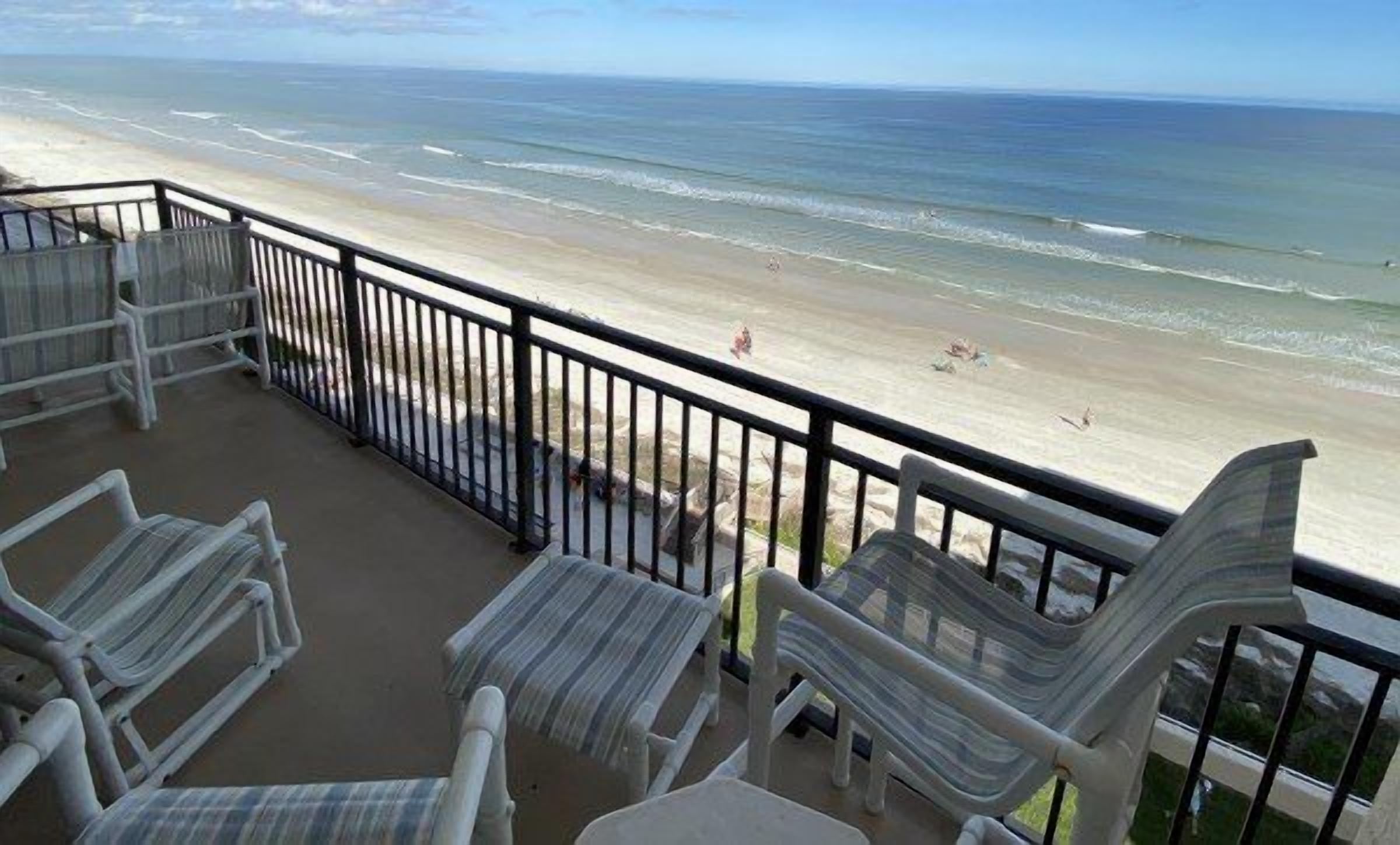 A balcony with a view of the beach, white chairs, and a railing.