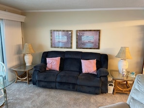 A living room with a dark sofa, two pink cushions, a glass coffee table, two side tables with lamps, and two framed pictures on the wall. - #705 Sandcastle 2/2 Oceanfront CondoTraffic Free Beach (Ponce Inlet)
