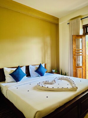 A hotel room with a bed, pillows, a wooden headboard, a window with curtains, and a wooden door.