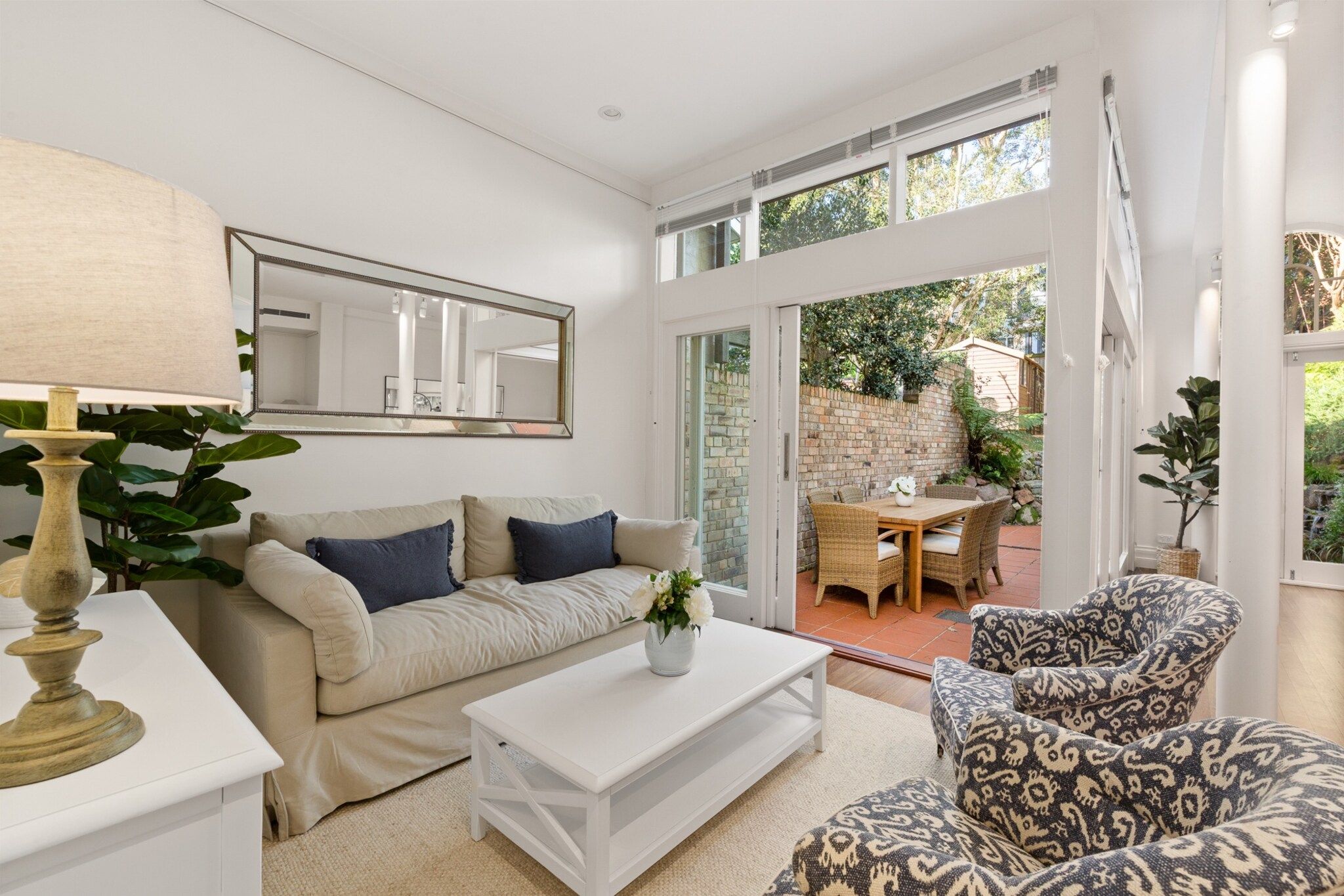 A modern living room with a sofa, coffee table, and patterned armchair. There is a dining area with a table and chairs outside.