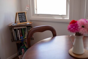 A room with a wooden chair, a small table, a bookshelf with books, a vase with flowers, and a window with natural light. - Charming 1-Bed Cottage in Historic Berrima (Berrima)