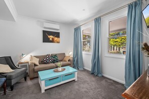 A living room with a sofa, a coffee table, a TV, and a window with curtains. - Charming 1-Bed Cottage in Historic Berrima (Berrima)