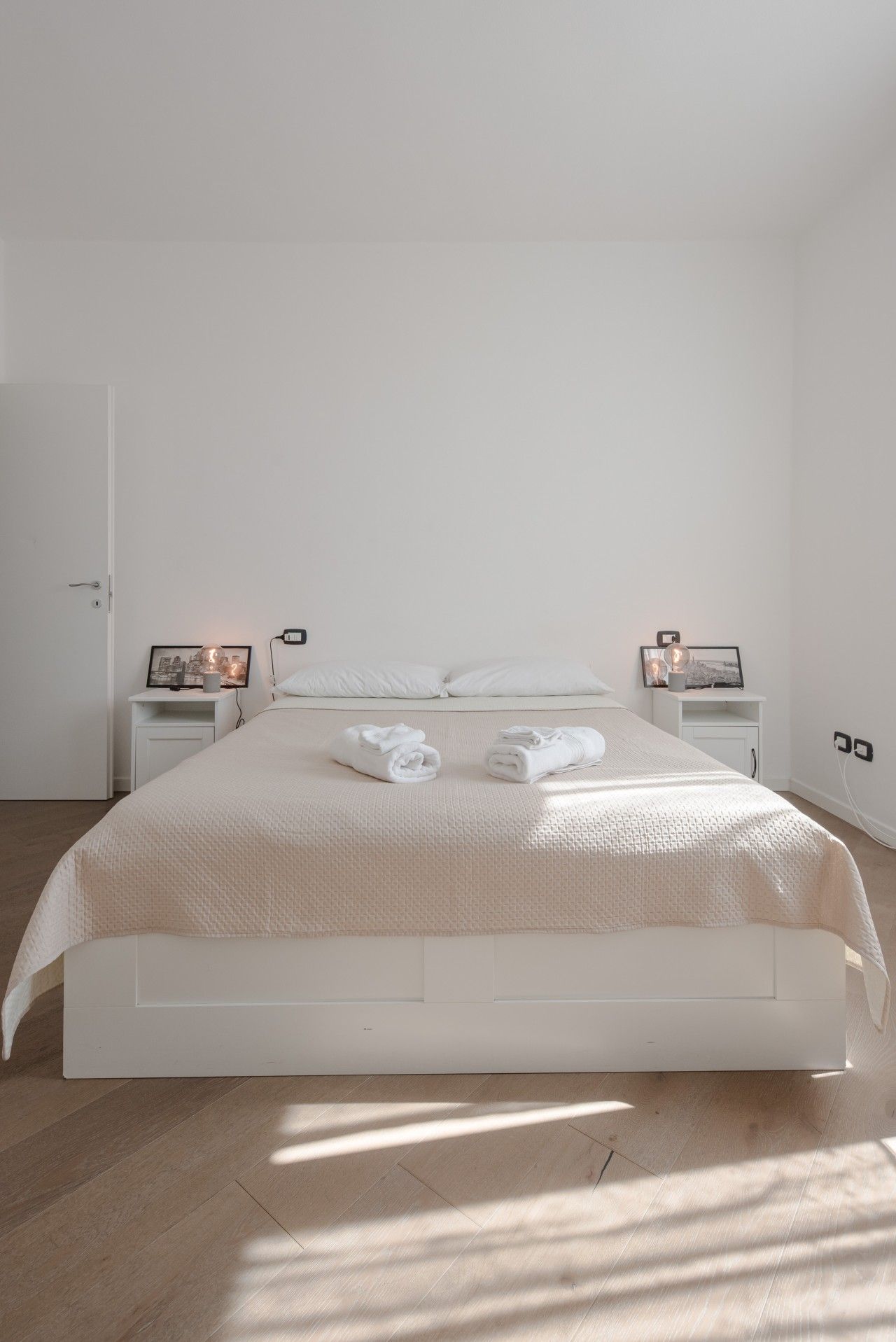 Een minimalistische slaapkamer met een groot bed, twee nachtkastjes met lampen en een deur.