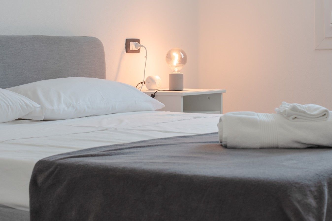 A neatly made bed with white bedding and a grey blanket, a bedside lamp, and a folded towel on the nightstand.