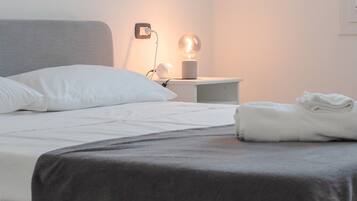 A neatly made bed with white bedding and a grey blanket, a bedside lamp, and a folded towel on the nightstand.
