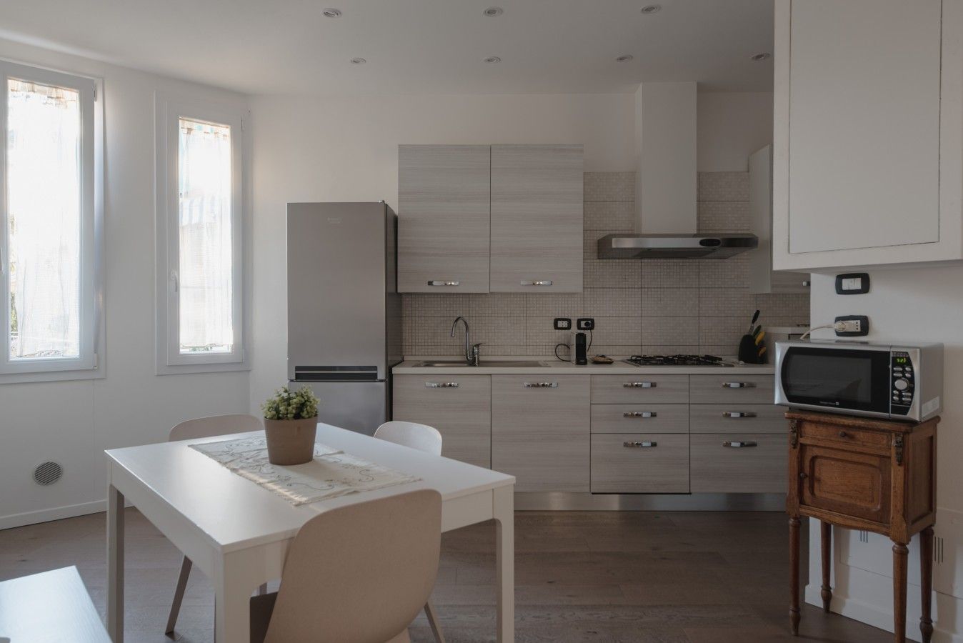 A modern kitchen with a dining area, featuring a white table, chairs, a potted plant, and a microwave on a wooden stand.