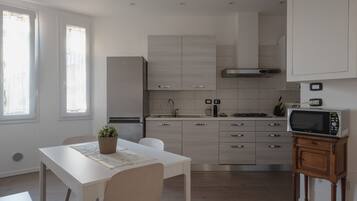 A modern kitchen with a dining area, featuring a white table, chairs, a potted plant, and a microwave on a wooden stand.