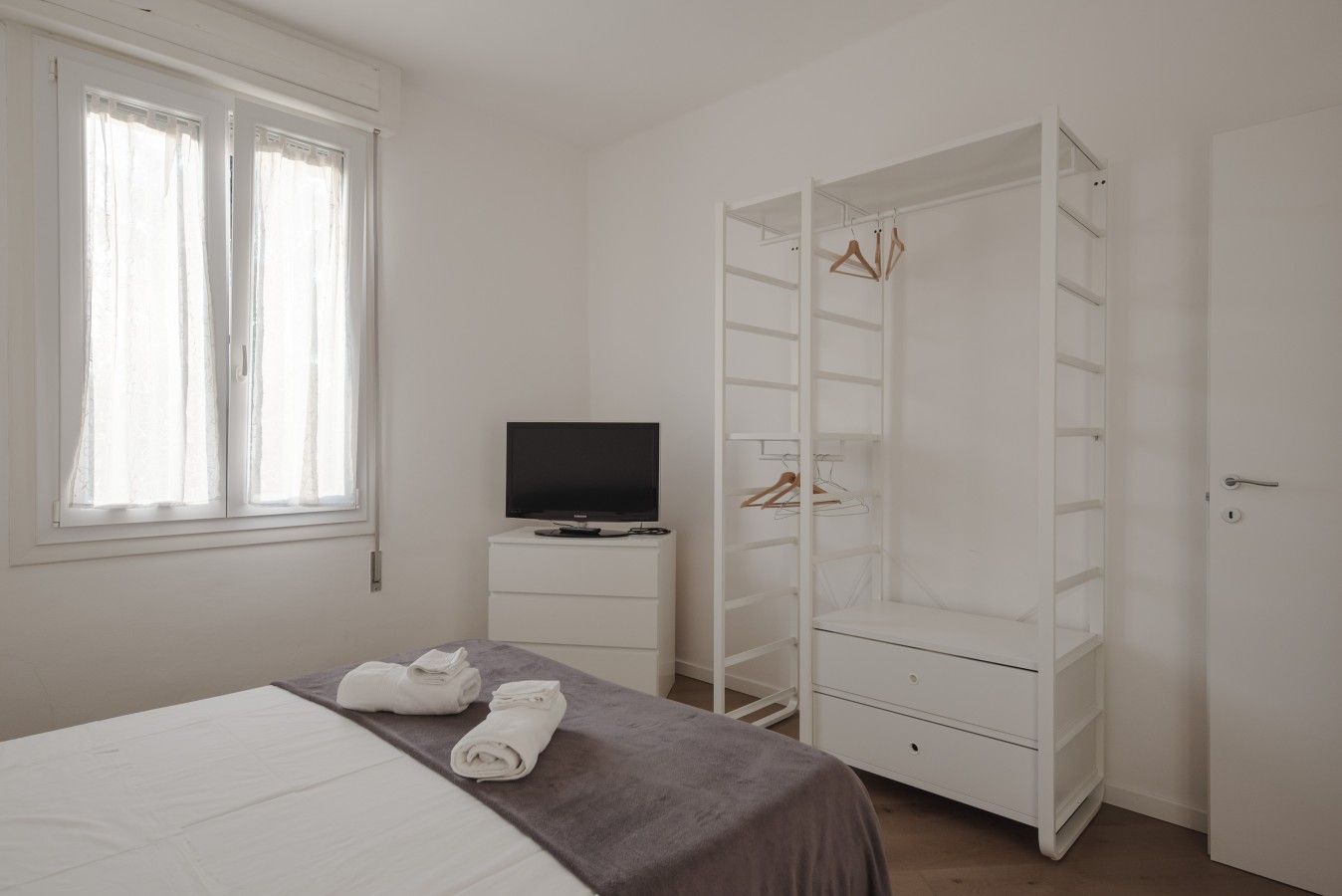 Quarto com cama, televisão sobre suporte, guarda-roupa com espaço para pendurar roupas e janela com cortinas.