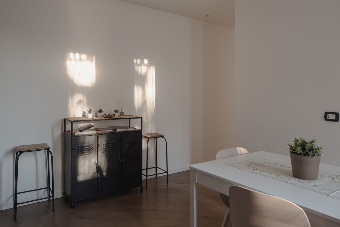 Ein schlicht eingerichteter Raum mit einem weißen Tisch, zwei Hocker, einer schwarzen Metallschrank und einer Topfpflanze.