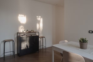 Ein schlicht eingerichteter Raum mit einem weißen Tisch, zwei Hocker, einer schwarzen Metallschrank und einer Topfpflanze.