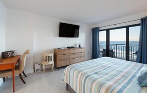 A hotel room with a bed, a TV mounted on the wall, a wooden dresser, a small side table, a chair, and a view of the ocean through large windows. - 711 Sandcastle N in Ponce Inlet, FL (Ponce Inlet)