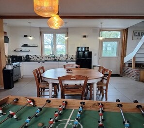A dining area with a round table and chairs, a kitchen with appliances and a refrigerator, and a foosball table in the foreground. - 3-Bedroom Country Cottage at Chez Violette (STEENBECQUE)
