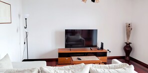 A living room with a flat-screen TV on a wooden stand, a floor lamp, a vase with dried plants, and a sofa with cushions. - Vale Formoso Funchal by Rentallido (Funchal)