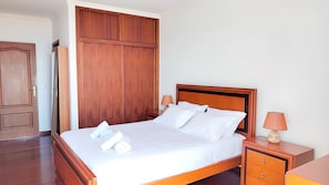 A neatly made bed with white linens and pillows, a wooden headboard, two bedside tables with lamps, and a wooden wardrobe. - Vale Formoso Funchal by Rentallido (Funchal)