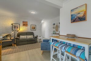 A cozy living room with a sofa, a dining table with benches, and a lamp. - Apollo Holiday Units - 2-Bed Unit in Narooma (Narooma)