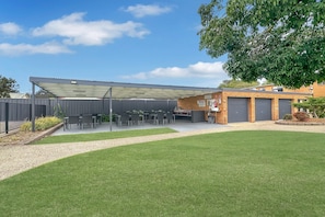 A covered outdoor seating area with tables and chairs, a grassy lawn, and a building with a garage door. - Apollo Holiday Units - 2-Bed Unit in Narooma (Narooma)