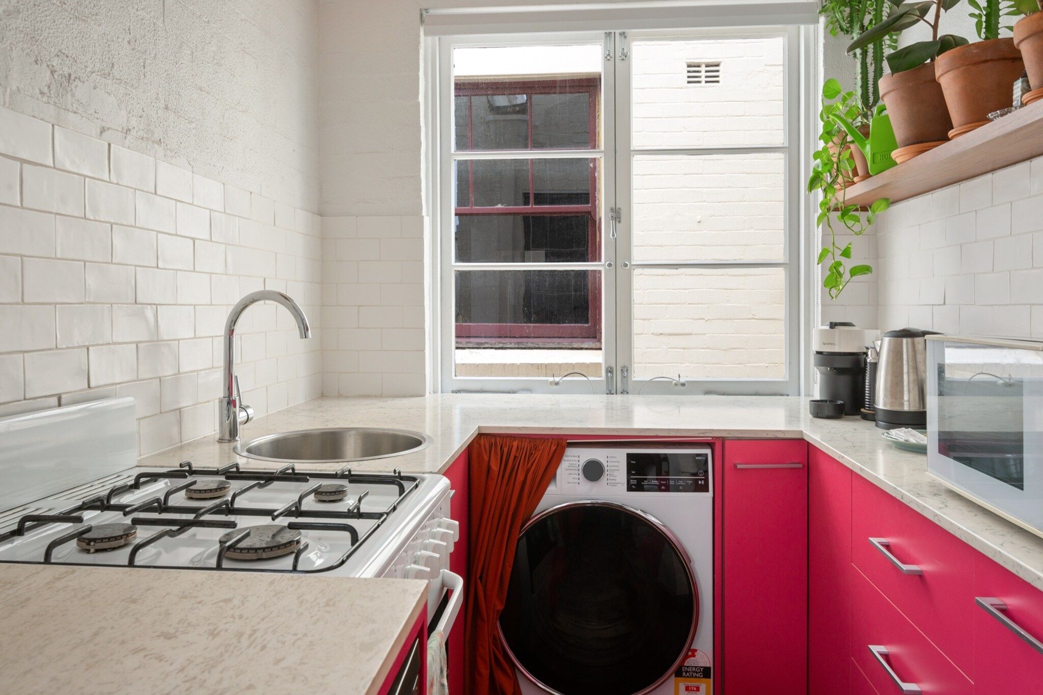 Een keuken met een gasfornuis, een spoelbak, een wasmachine en een raam met uitzicht op een wit gebouw.