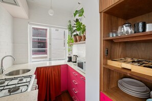 Eine Küche mit Gaskochfeld, Spüle und einem Fenster mit Vorhängen. Auf einem Regal stehen Blumentöpfe und ein Holzschrank mit Geschirr.
