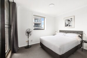 A hotel room with a bed, a standing fan, a window with a view, and a framed picture on the wall. - Spacious 2-Bed Apartment Near Parramatta CBD (Parramatta)