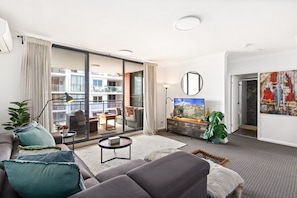 A modern living room with a sofa, coffee table, and a TV. There is a balcony with a view of a building. - Spacious 2-Bed Apartment Near Parramatta CBD (Parramatta)