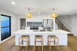 A modern kitchen with a central island, stainless steel appliances, and a staircase leading to an upper level. - Coastal Pool 3-Bed Townhouse Near Broulee Beach (Broulee)