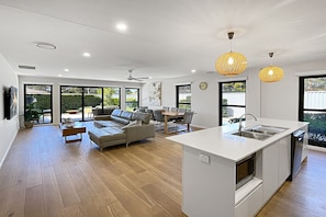 A modern kitchen with a central island, a dining area with a sofa, and large windows offering views of the outdoors. - Coastal Pool 3-Bed Townhouse Near Broulee Beach (Broulee)