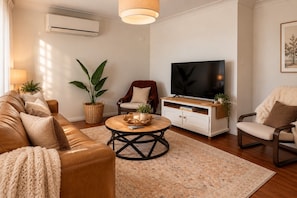 A living room with a leather sofa, a coffee table, a TV on a white cabinet, and a large plant. - Garden 2-Bed Villa in the Heart of Bowral (Bowral)