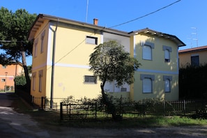 A two-story building with a yellow facade, a visible window with a metal grille, and a tree in front. - Comfy home near the beach (Rosolina Mare)