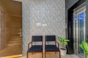 Um corredor com papel de parede estampado, duas cadeiras de madeira com almofadas pretas, uma planta em vaso e uma porta com design geométrico.