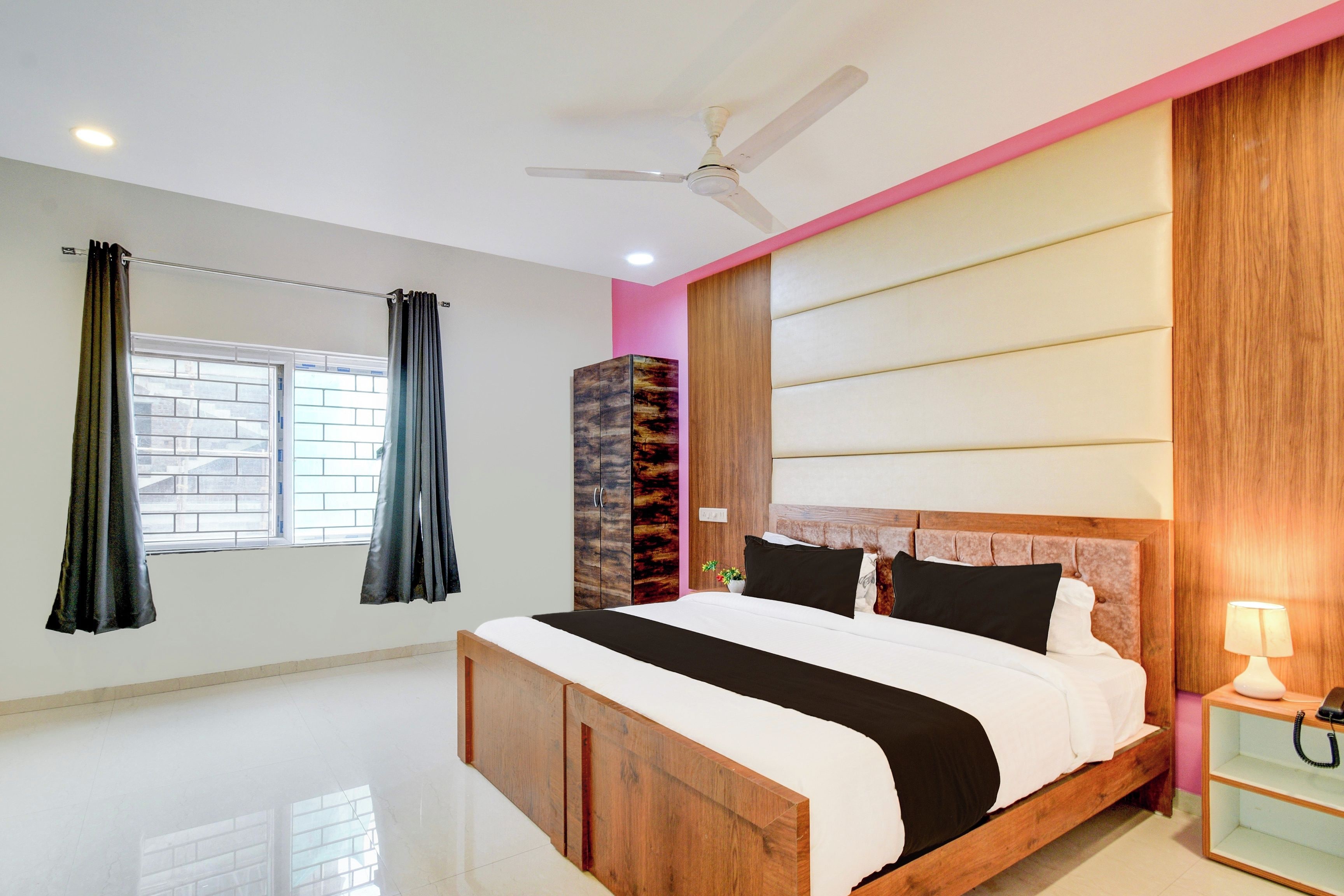 Un dormitorio con cama, ventilador de techo, ventana con cortinas y mesita de noche con lámpara.