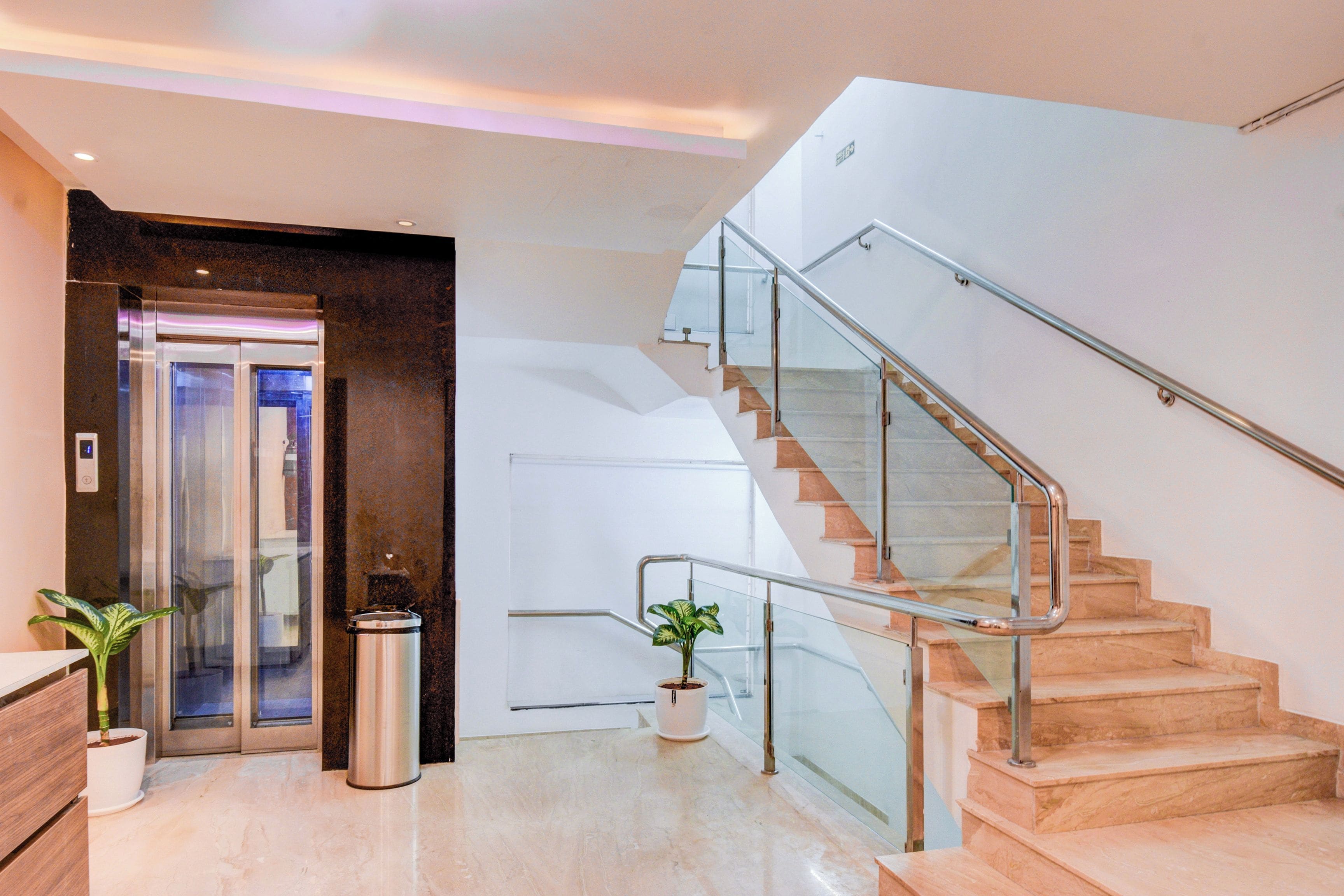 A modern hotel lobby with a glass elevator, a staircase with metal railings, and a potted plant.