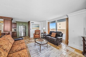A living room with a sofa, a coffee table, and a leather chair. There is a kitchen in the background with a refrigerator and cabinets. - #801 Blue Surf  Luxury 3/3 Oceanfront Condo Traffic Free Beach (Daytona Beach Shores)