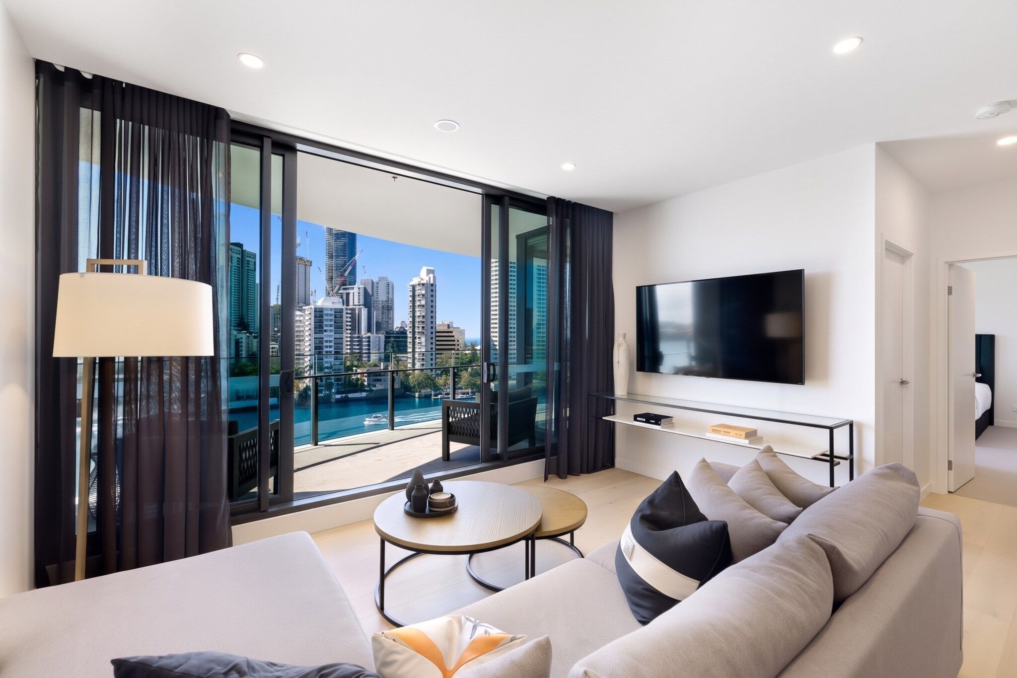 A modern living room with a large TV, comfortable seating, a small table, and a view of a city skyline and water.