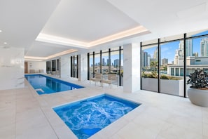 A modern indoor pool area with a city view, a separate hot tub, and seating. - Luxury Chevron Island Retreat | Water & City Views (Surfers Paradise)