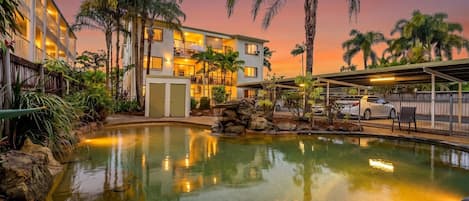 A hotel with a swimming pool, palm trees, and a sunset sky.