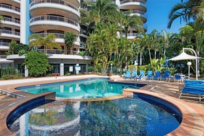 A swimming pool with a curved design, surrounded by lounge chairs and palm trees, with a multi-story building in the background. - High-Rise 2-Bed Apartment in Surfers Paradise (Surfers Paradise)