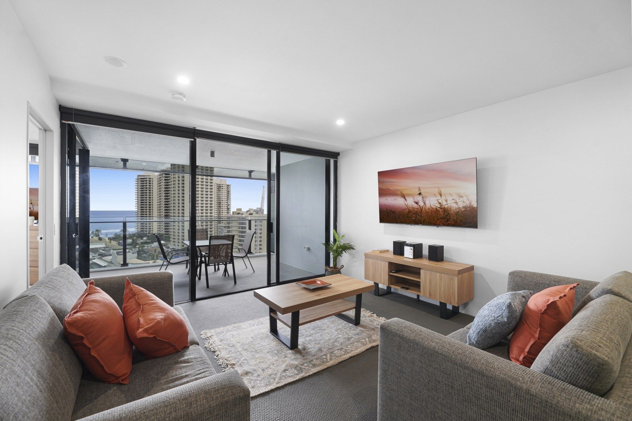 A modern living room with a sofa, coffee table, and TV. There is a balcony with a view of tall buildings.