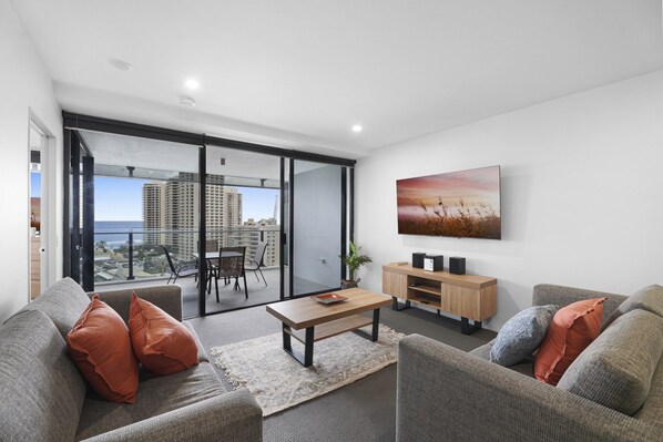 Ein modernes Wohnzimmer mit einer Couch, einem Couchtisch und einem Fernseher. Ein Balkon bietet Blick auf Gebäude und die Stadtlandschaft.
