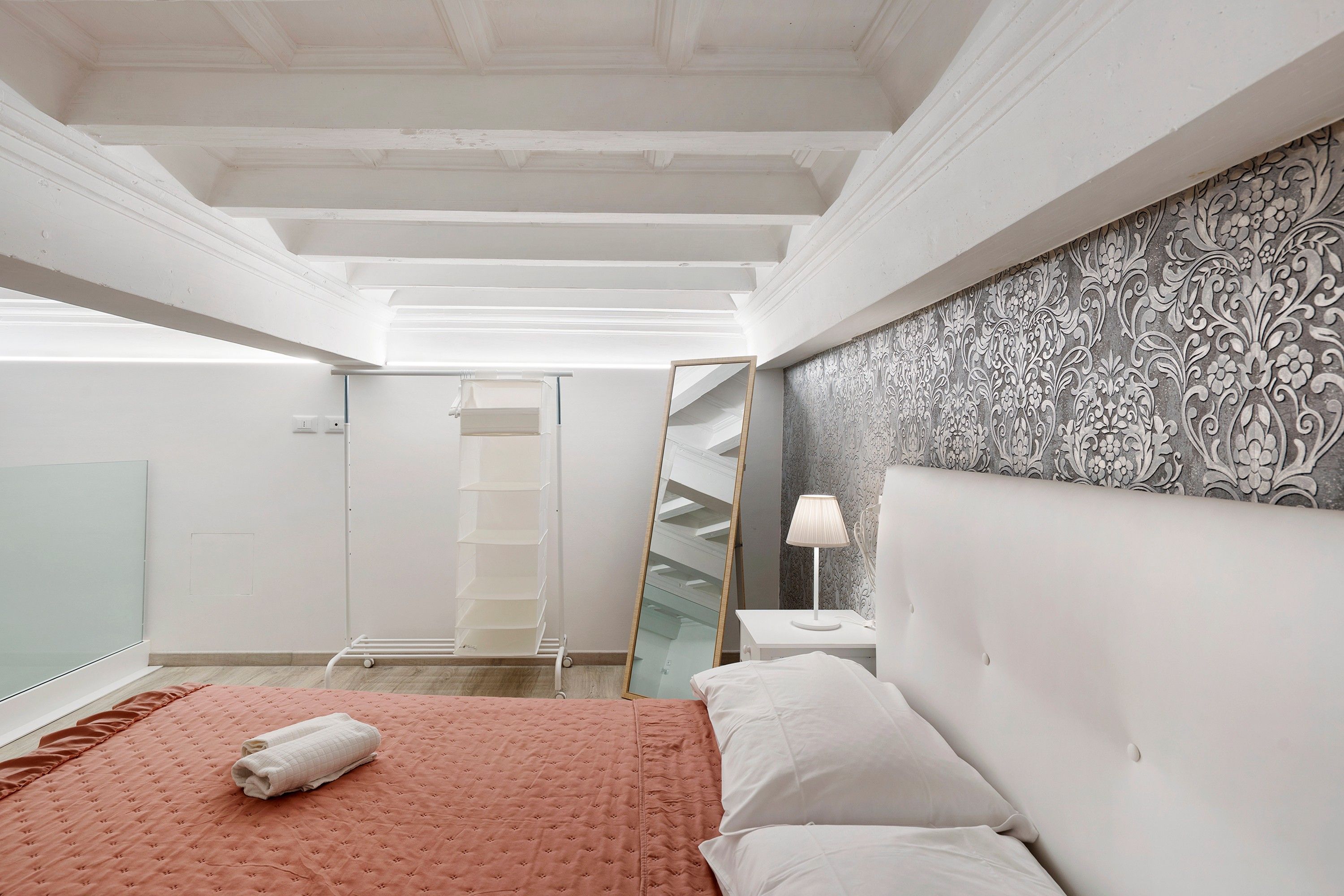 A bedroom with a bed, a bedside table, a lamp, a mirror, and a patterned wall.