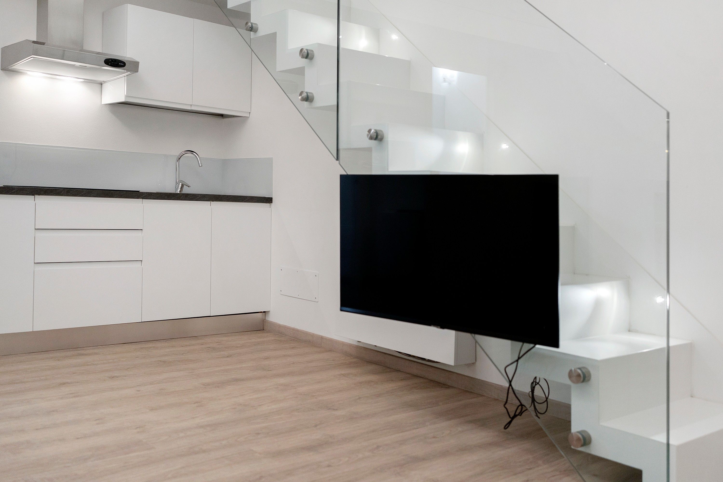 A modern kitchen with a built-in stove, a flat-screen TV mounted on a glass wall, and a wooden floor.