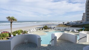 Área de piscina com vista para a praia e oceano, rodeada por espreguiçadeiras e uma palmeira.