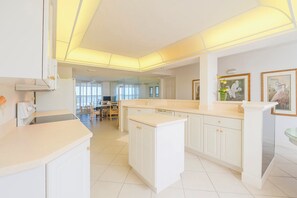 A modern kitchen with white cabinetry, a central island, and a dining area in the background. - 8505 Towers in Ponce Inlet, FL (Ponce Inlet)