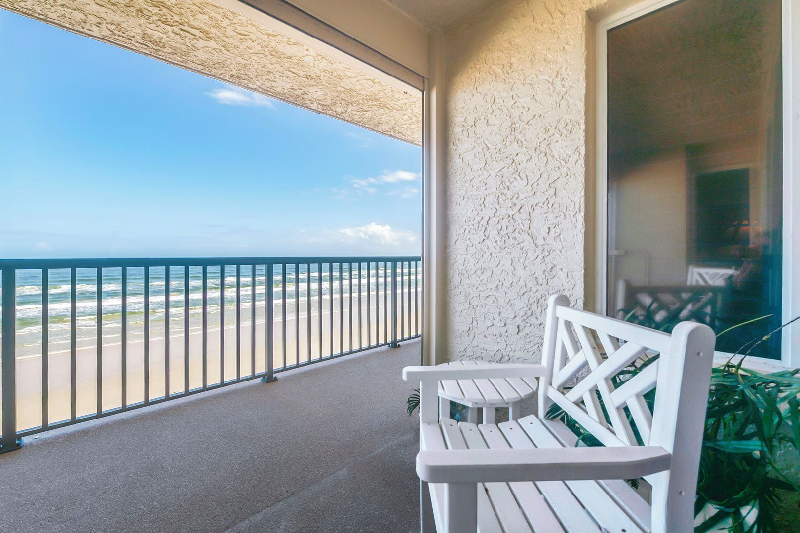 A balcony with a view of the beach and ocean, two white chairs, and a glass door.