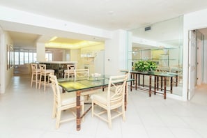 Uma sala de jantar moderna com mesa de vidro e cadeiras de vime, cozinha ao fundo, e divisória de vidro.