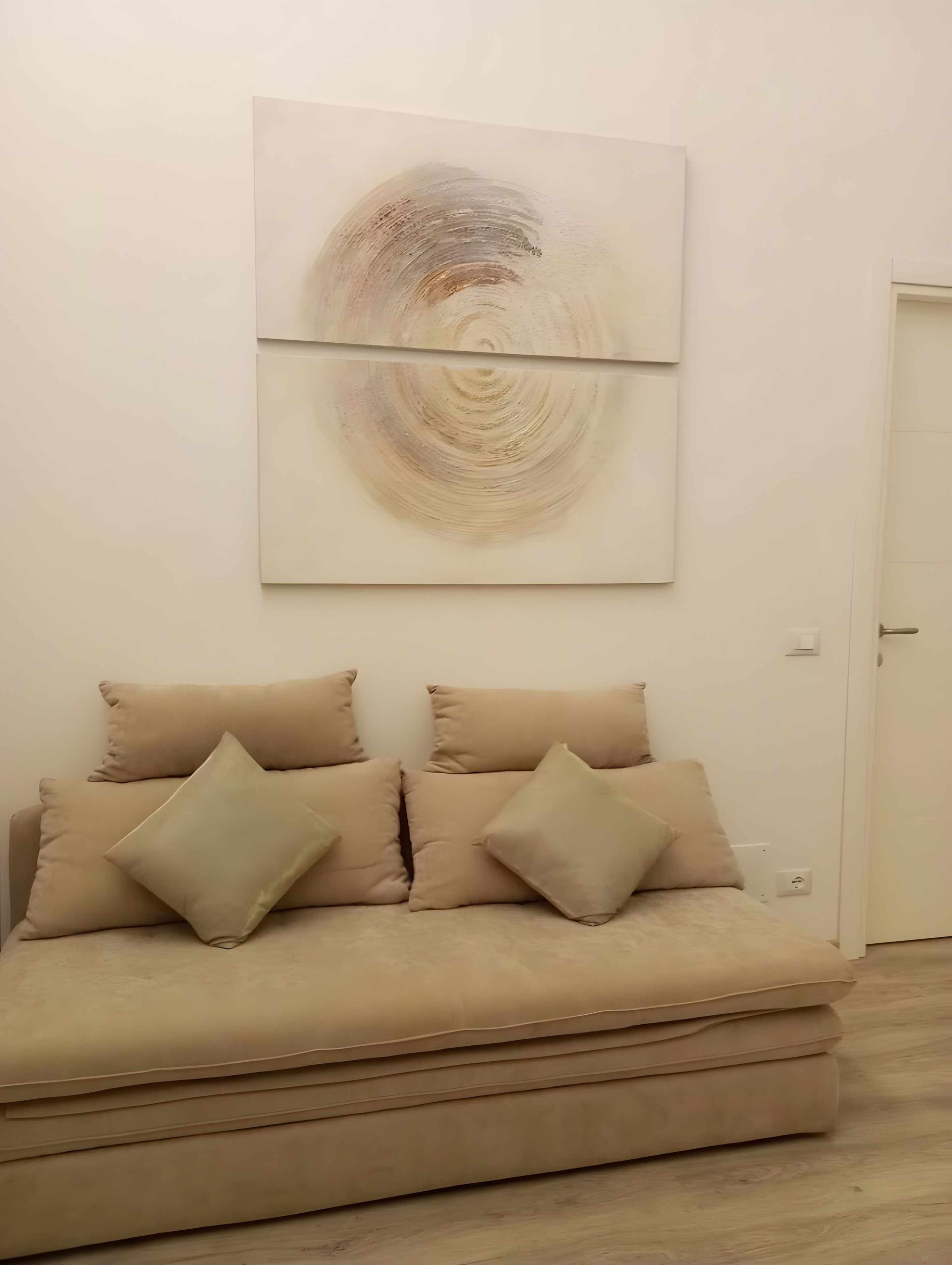 Une pièce avec un canapé beige, deux coussins et un grand tableau abstrait accroché au mur.
