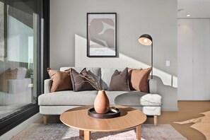 A modern living room with a grey sofa, brown cushions, a round wooden coffee table, and a framed abstract art piece on the wall. - High-Rise 2-Bed Apartment Near Southbank (South Melbourne)