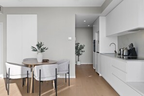 A modern kitchen with white cabinetry, a dining area with a round table and two chairs, and a potted plant. - High-Rise 2-Bed Apartment Near Southbank (South Melbourne)