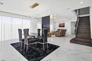 Un salon moderne avec un coin repas, comprenant un ensemble de salle à manger noir et doré, un grand téléviseur et un escalier.
